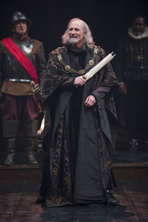 Lear wears black robes with purple pattern, a chain with several medallions draped over his shoulders, and long silver hair to his shoulders, and he carries a scroll in his left hand: a soldier in Renaissance breastplate armor and helmet with knee boots and a red sash stands behind on the left, and on the right is a lord in ruff collar